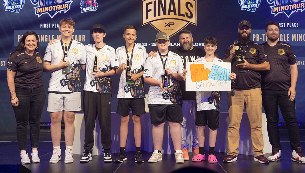 A group of players and coaches in esports jerseys on stage, one player holds a sign that reads “For Hank.”