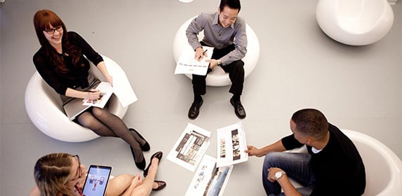 Four students sit together on round, white chairs and review printed slides of marketing graphics.