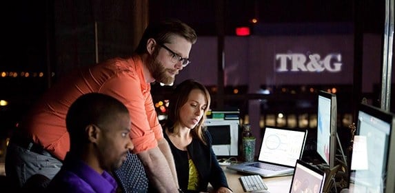 An instructor leans in to review and analyze data compiled on multiple monitors with two seated students.