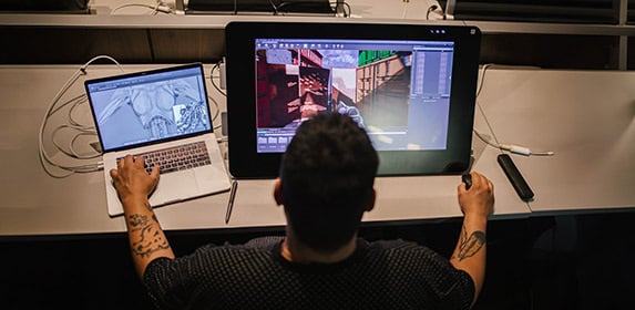 Students listen intently to a lecture with MacBooks, pen tablets, and sketch pads arrayed across a table.