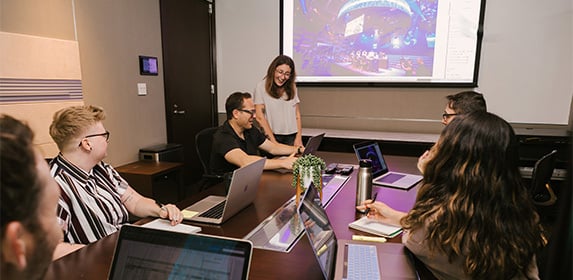 A group of five people are seated at a table in a conference room while a woman in the front of the room looks on smiling and pointing at a seated attendee's laptop.