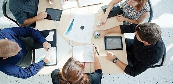 Seated at a large wooden table, multiple students discuss a printed marketing graph while taking notes on journals, tablets, and legal pads.