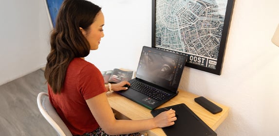 A Game Art student sits at a desk. They are using a tablet to draw a 3D-rendered face, which is displayed on their laptop.