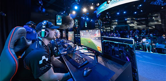 A man at an esports event playing on a computer while sitting in a gaming chair with an onlooker behind him.