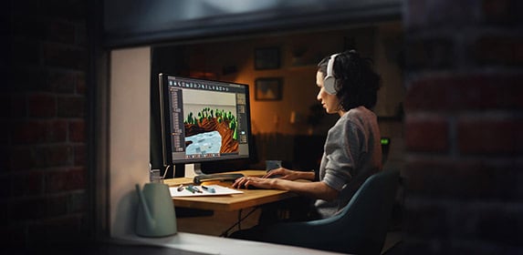 A Game Design student wears headphones and sits at a desk. They are working at a desktop computer with game design software on the screen.