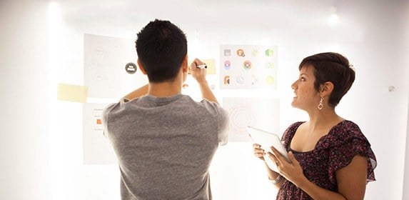 A student hangs a series of logos on a board while another student compares with their tablet.