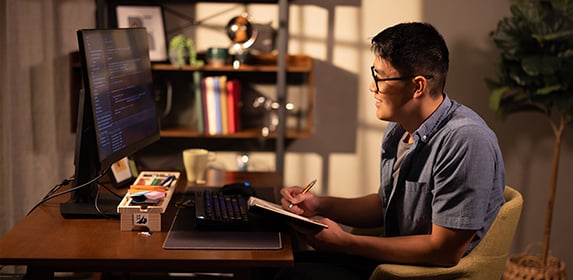 A smiling student holding a pen and notebook works at a desktop computer.