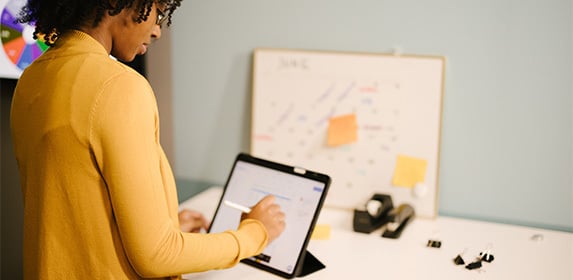 A woman in a yellow blouse is writing on a tablet with a smart pen while at a standing desk with her back to the camera.