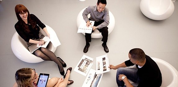 Four students sit together on round, white chairs and review printed slides of marketing graphics.