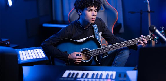 A music production student is playing an acoustic-electric bass guitar while seated in front of a MIDI keyboard and launchpad from their Project LaunchBox™.