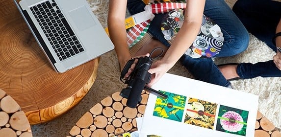 A student surrounded by storyboards and props uses a digital camera to record a video.