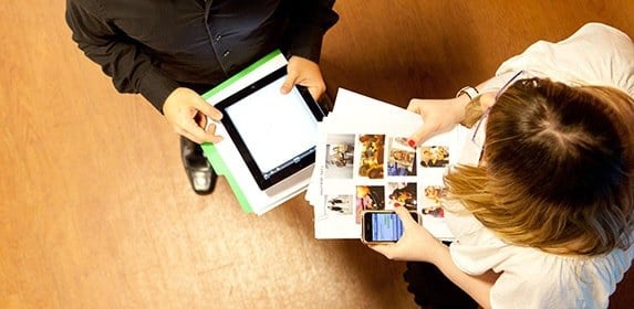 Two students holding printed documents with photos of a film set talk to one another in a hallway.