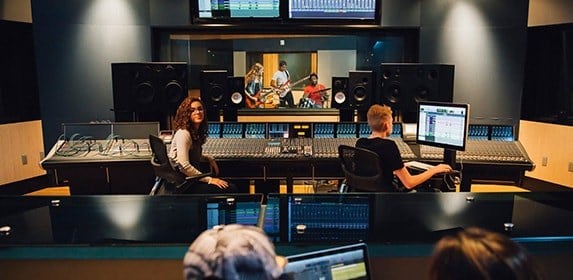 A group of students monitors audio levels on a massive soundboard for a band playing in a recording studio classroom.