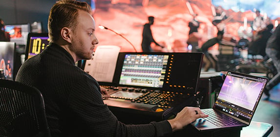A student works in front of an audio console and a laptop in a virtual production studio.