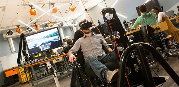 A student wearing a virtual reality headset tests a racing simulator in a special rig designed to simulate a driver’s seat.