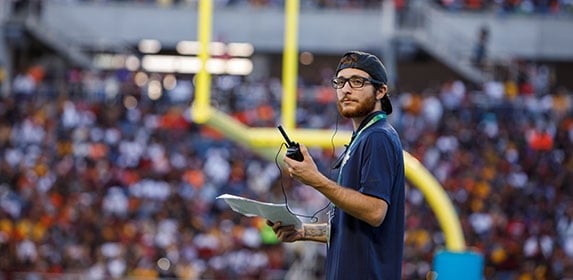 A student holding a walkie-talkie and loose documents stands across from a field goal post.