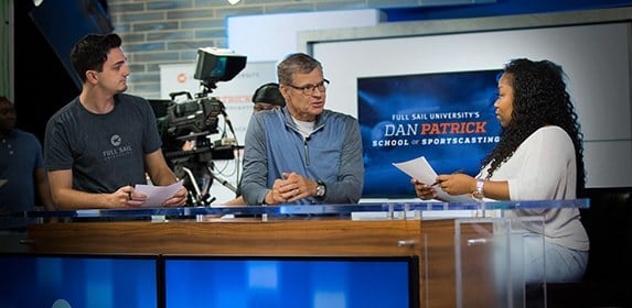 Sportscasting icon Dan Patrick gives advice to students at a broadcast desk before they go live.