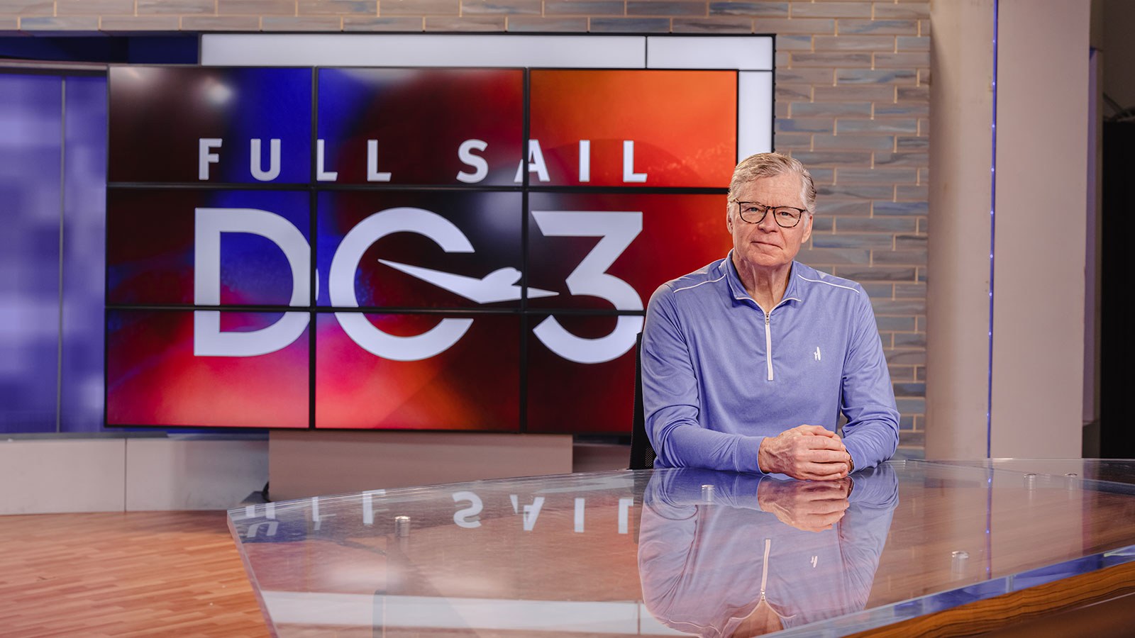 Photograph of Dan Patrick sitting at an anchor desk in front of a screen with Full Sail DC3 logo displayed behind him.