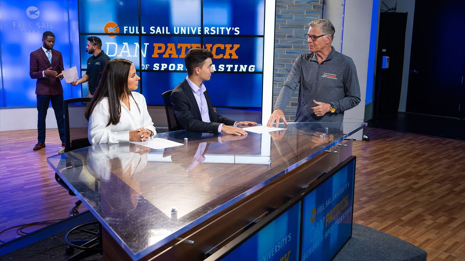 Sportscaster Dan Patrick standing at a news desk speaking with two students at the Full Sail University Dan Patrick School of Sportscasting soundstage.