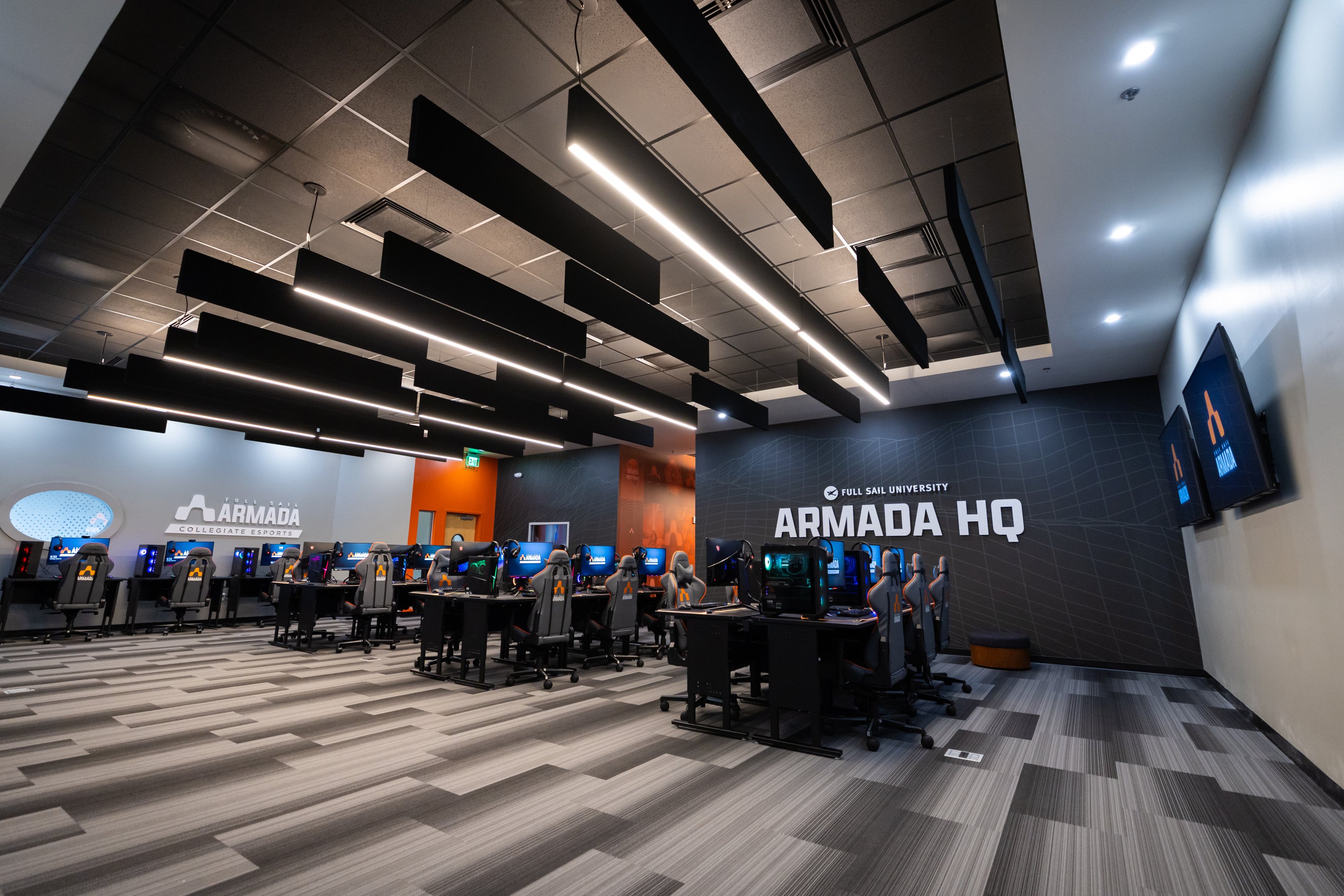 A room with gray walls and carpeting. The word Armada is written across the wall in white and several stations with gaming PCs are against the wall.