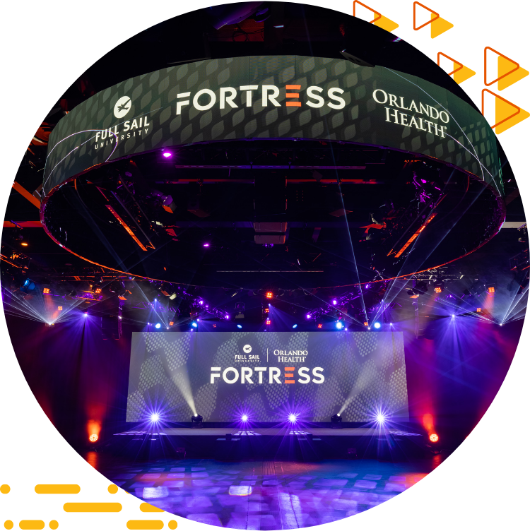A photograph of the Full Sail Orlando Health Fortress, with spotlights and multiple display screens containing logos for the esports space.