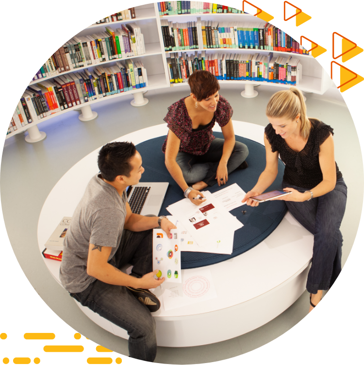 Aerial photo of three students collaborating on design work while seated on a large ottoman, books are in the background.